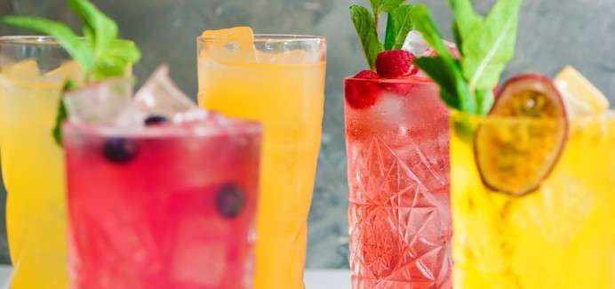 Colorful iced drinks sit on a table, garnished with mint, raspberries, blueberries, and passion fruit. Multiple tall glasses hold pink, yellow, and orange beverages, against a blurred gray background.