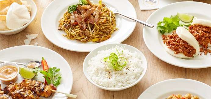 Assorted Asian dishes are served: stir-fried noodles with meat, steamed rice, bao buns with shredded meat, chicken satay with peanut sauce, lime wedges, cilantro, prawn crackers on a wooden table.