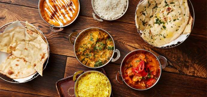 Indian dishes sit arranged on a wooden table: curries in metal bowls, yellow rice, plain rice, naan breads, and a creamy orange curry, garnished with herbs, ready for serving.