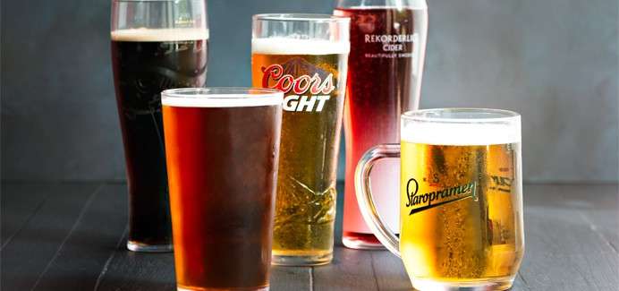 Assorted beer and cider glasses stand filled on a dark tabletop; labels read Coors Light, REKORDERLIG CIDER, and Staropramen, with amber, dark, and red drinks against a gray backdrop.