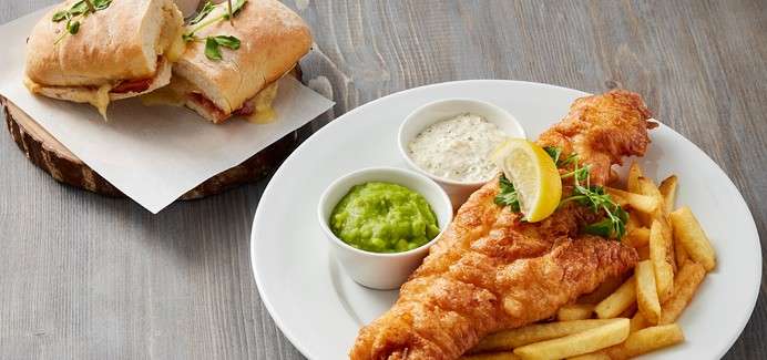 Fried fish rests on fries, garnished with lemon and herbs, accompanied by bowls of tartar sauce and mushy peas; a toasted sandwich sits on parchment atop a wooden board.