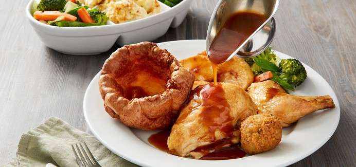 Roast chicken and sides receive gravy from a metal jug, on a white plate with Yorkshire pudding, potatoes, stuffing ball, and vegetables; side bowl and cutlery rest on a wooden table.