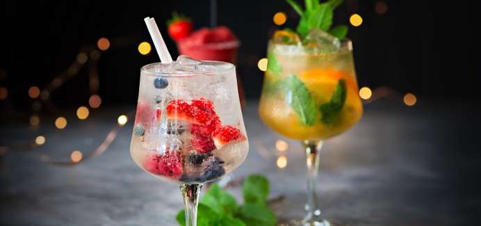 Two garnished cocktails sit on a table: a berry-filled spritzer with straw and a mint-citrus drink with ice, while blurred fairy lights twinkle in a dark, festive background.