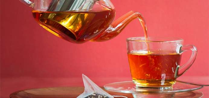 Glass teapot pours amber tea into a clear cup on a saucer. Nearby, a pyramid tea bag rests on a wooden board. Scene set against a solid red backdrop.