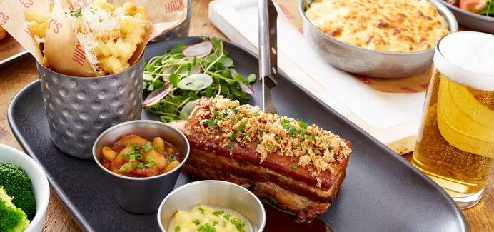 Pork belly sits crumb-topped in sauce; accompanied by fries in a metal cup, salad greens, two dipping sauces, broccoli, a cheesy bake, and a frosty beer on a wooden table.