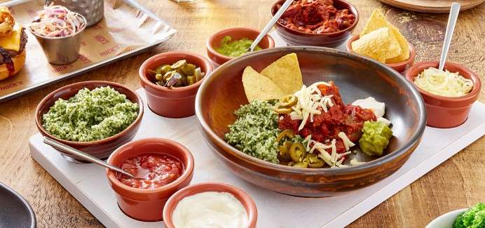 Bowl of nachos topped with salsa, shredded cheese, guacamole, jalapeños, and chips sits central, surrounded by small bowls of dips and condiments on a wooden table.