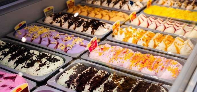 Gelato trays sit in a refrigerated display, swirled and topped with chocolate, sprinkles, and fruit sauces, while small flavor tags stand upright, arranged in colorful rows inside an ice-cream shop.