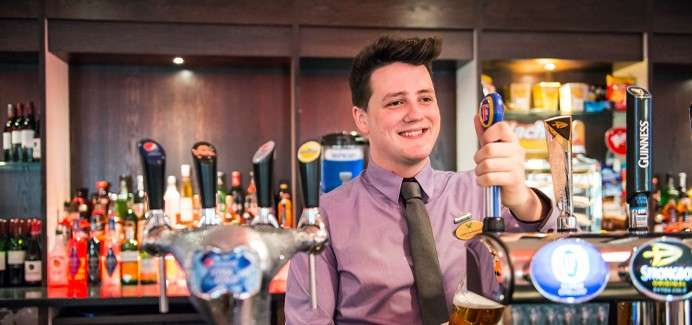 Bartender pulls a tap to pour beer; behind him, shelves of liquor and illuminated dispensers in a bar setting. Text: GUINNESS.