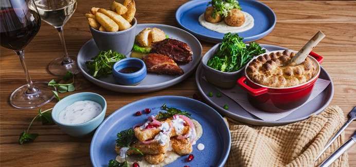 Plated dishes sit arranged on a wooden table: a red pot pie, ribs, potato wedges, croquettes with sauce, greens, and dipping bowls, accompanied by red and white wine glasses.