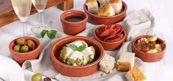 Tapas bowls display soft cheese with pesto, olives, chorizo slices, bread cubes, and a dark dip, sitting on a linen cloth, with two sparkling wine glasses on a rustic white tabletop.