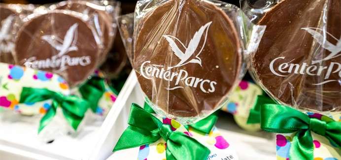 Chocolate lollipops display “CenterParcs” with a bird logo, wrapped in clear plastic and tied with green bows, arranged on a shop shelf with colorful polka-dot packaging.