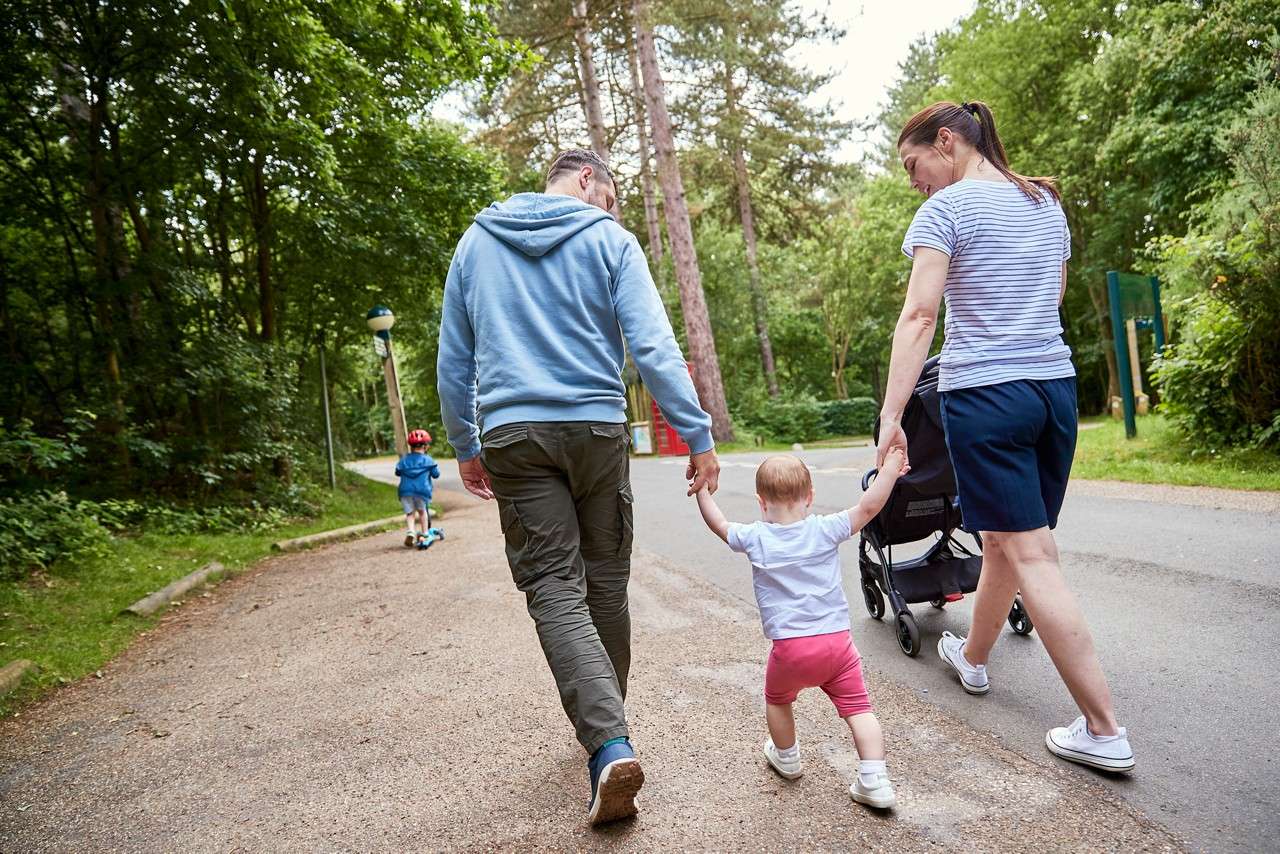 Toddler walks while holding two adults’ hands, beside a stroller, on a paved path. Another child rides a scooter ahead. Surrounding trees and greenery line the quiet park road.