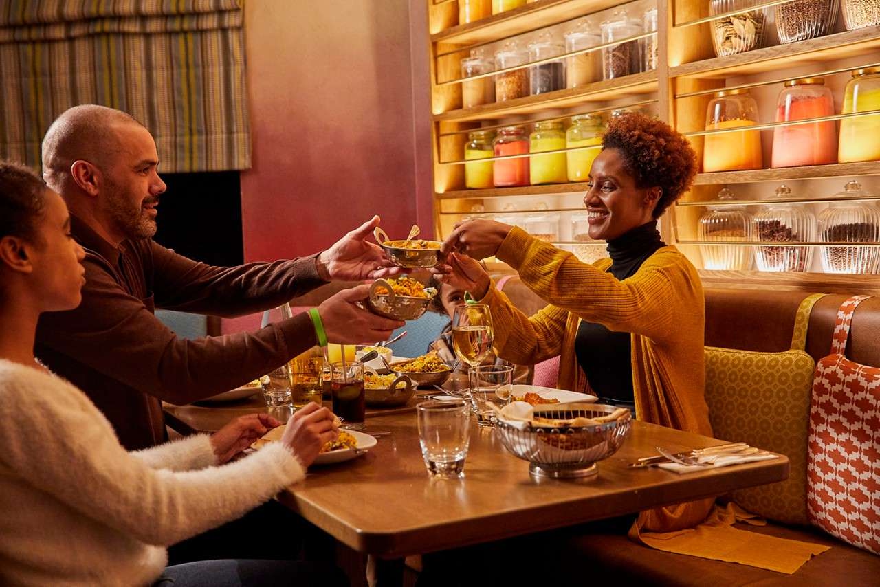 Child in a yellow life jacket laughs as an adult lifts them above water; another adult smiles nearby in an outdoor pool bordered by lounge chairs, railing, and tall trees.