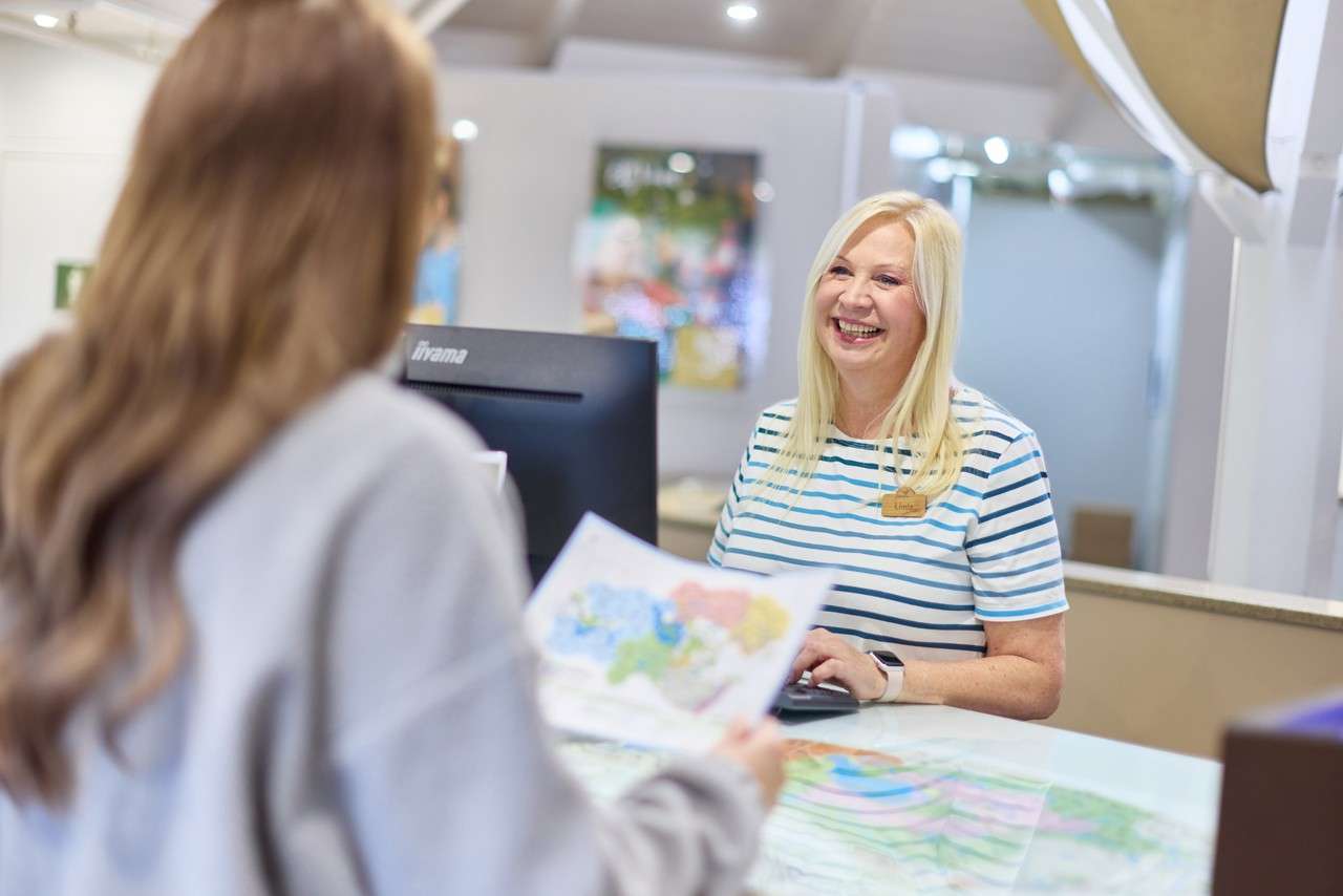 Child in a yellow life jacket laughs as an adult lifts them above water; another adult smiles nearby in an outdoor pool bordered by lounge chairs, railing, and tall trees.