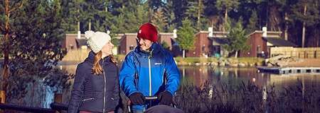 Two adults walk, one pushing a stroller, on a lakeside path; they wear winter hats and jackets; cabins and pine forest sit across the water in cool morning light.
