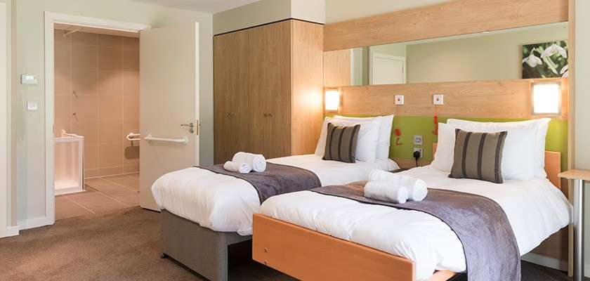 Two twin beds with folded towels rest side by side, lamps lit, facing a long mirror headboard; a modern hotel room with wardrobe, carpet, and an open bathroom doorway.