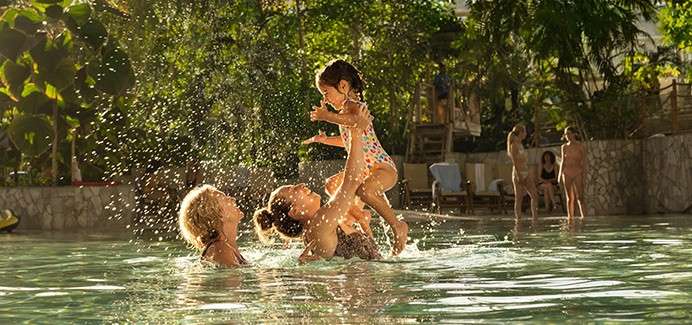 Child in a polka-dot swimsuit—being lifted laughing into the air—by two adults amid splashing water, in a sunlit, shallow pool; tropical plants, lounge chairs, and other swimmers in the background.