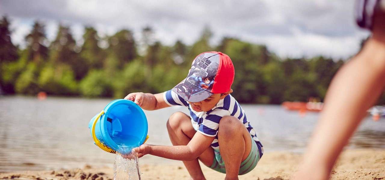 Child pours water from a blue bucket while crouching on sandy beach; wearing a striped shirt and cap, with a calm lake, distant trees, and cloudy sky in the background.