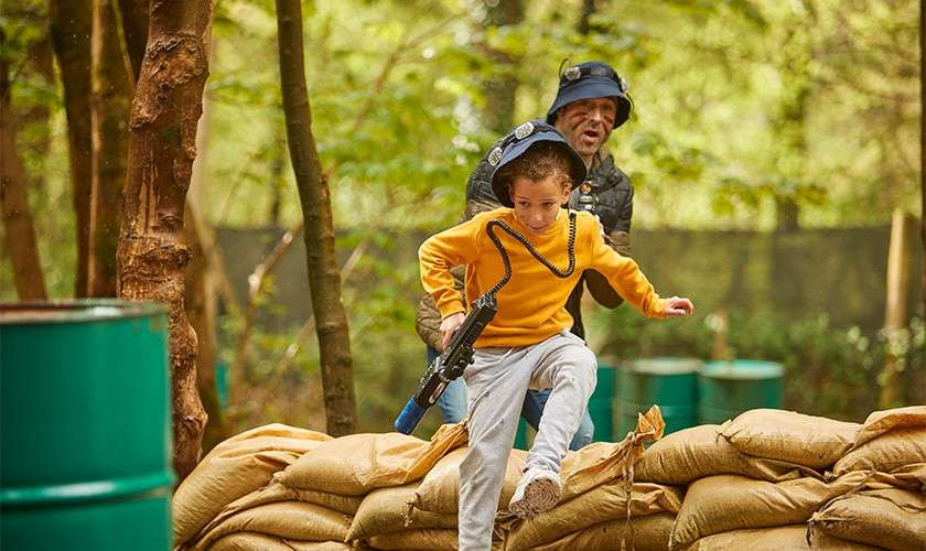 Child with a toy laser gun jumps over a sandbag barricade while an adult in protective gear follows, in a wooded outdoor play area with green barrels and trees.