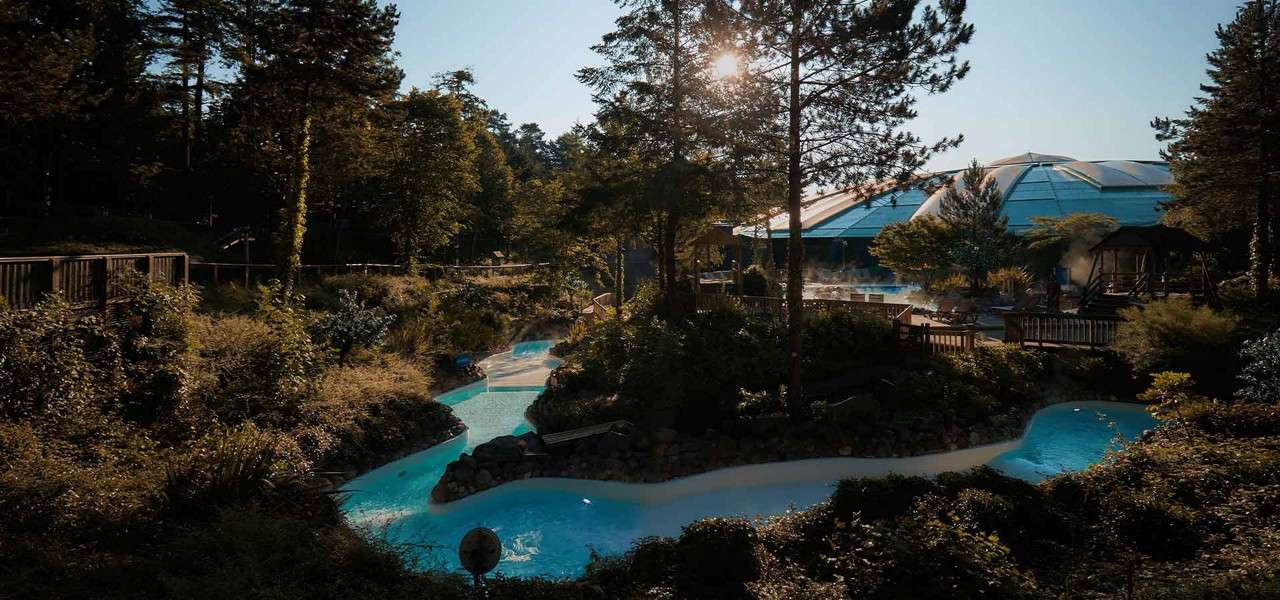 A turquoise lazy river winds gently through landscaped rocks and dense trees, steaming in cool air, beside wooden decks and a large domed water park building at sunset.