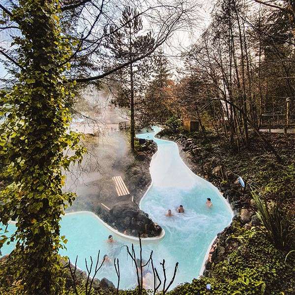 People soak and lounge in terraced turquoise hot spring pools, steam rising, surrounded by rocks, trees, and dense greenery; the pools wind through a forested setting with benches nearby.