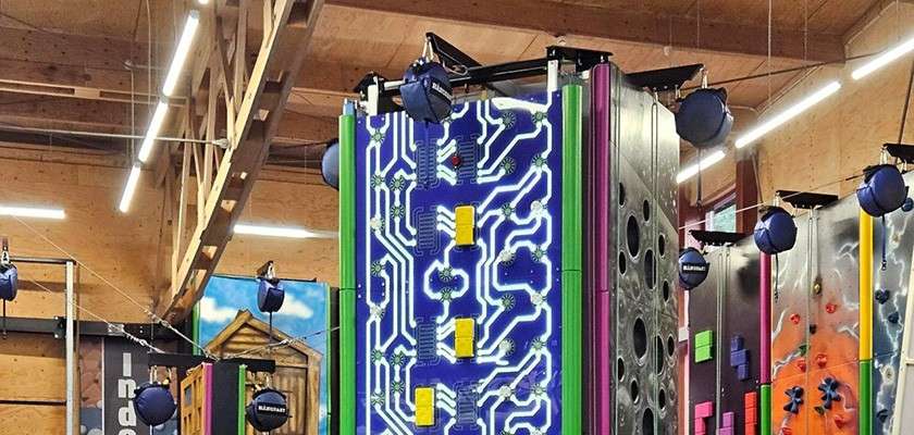 Colorful climbing walls rise vertically, fitted with auto-belay reels, within an indoor gym featuring wooden rafters, bright strip lights, and themed panels with varied holds and textures.