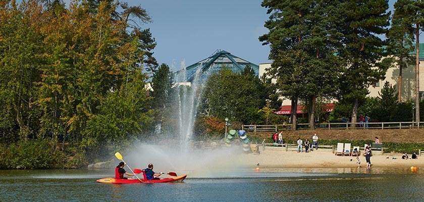 Red tandem kayak paddles across a calm lake, passing a tall fountain; nearby, people relax on a sandy beach beneath trees, with buildings and a glass-roofed structure in the background.