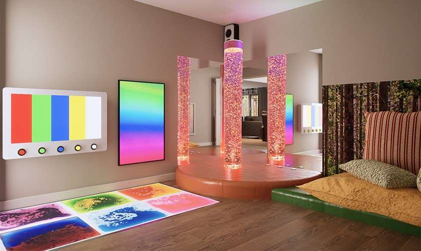 Three glowing bubble tubes shimmer on a round platform, illuminating a sensory room with color-changing wall panels, interactive light-up floor tiles, cushioned seating, and a forest-themed mural along one wall.