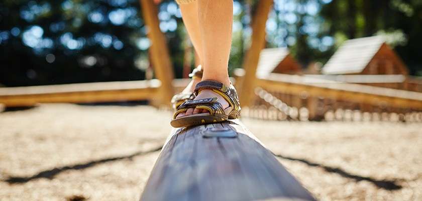 Sandaled feet balance on a narrow wooden beam, moving forward carefully in a playground. Sunlit timber structures and wood-chip ground surround, with blurred trees and cabins in the background.