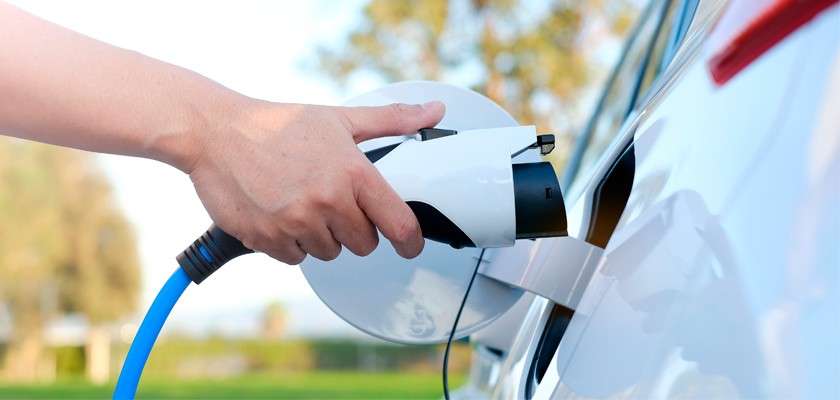Charging connector plugs into a white electric car’s port; a hand inserts the nozzle. Context: parked outdoors in daylight, blurred greenery and sky in the background.