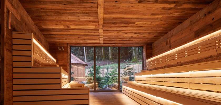 Wooden sauna benches glow with recessed lighting, lining both sides. A glass door frames evergreen trees and a small hut outside, within timber walls and ceiling creating a warm space.