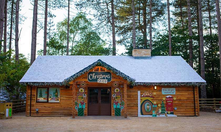Wooden shop displays festive decorations under a snowy roof; sign reads “Christmas SHOP.” A measuring board says “How do you measure up?” with “5'0"” marked. Surrounded by tall trees in a forest clearing.
