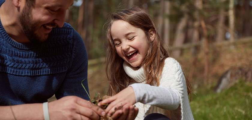 Smiling child laughs while handling leaves and twigs, guided by an adult beside her; both crouch on grass in a sunlit forest clearing with blurred trees in the background.