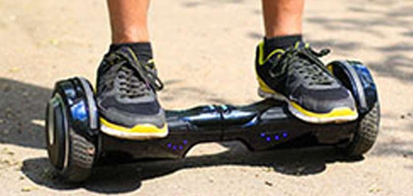Feet in black sneakers balance on a two-wheeled hoverboard, gliding along a sunlit, paved outdoor path.