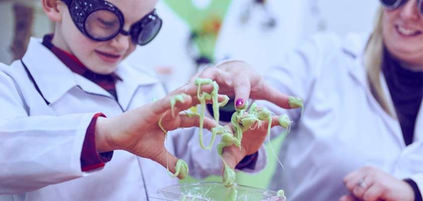 Child’s hands stretch green slime, dripping between fingers, over a clear container; supervised by an adult, both wearing lab coats and safety goggles in a playful classroom science setting.