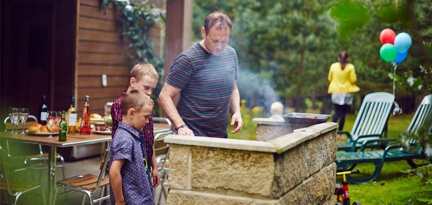 Man grills on a stone barbecue while two children watch, smoke rising; in a garden with lawn chairs, a table with food and drinks, balloons, and a few people mingling in the background.