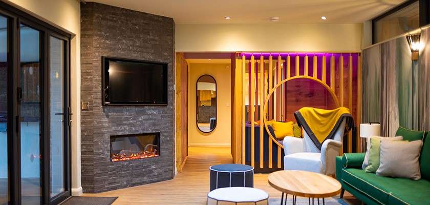 Living room shows seating, electric fireplace flickering, and wall-mounted TV; a circular wooden divider frames lounge chairs; sliding glass doors open to a pool area; warm lights illuminate the space.