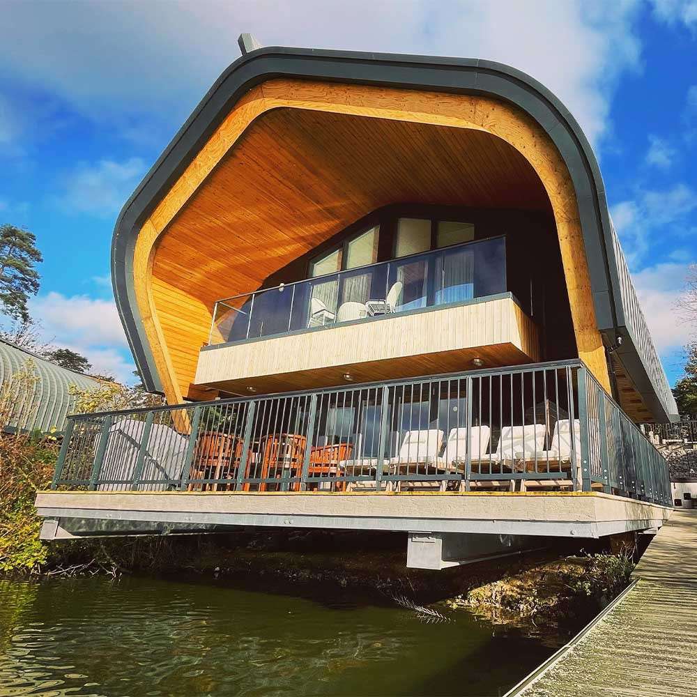Modern house with sweeping curved roof shelters glass-balustraded balcony and lower deck with lounge chairs, overhanging a calm waterway; wooden soffit gleams under blue sky, beside a dock and foliage.