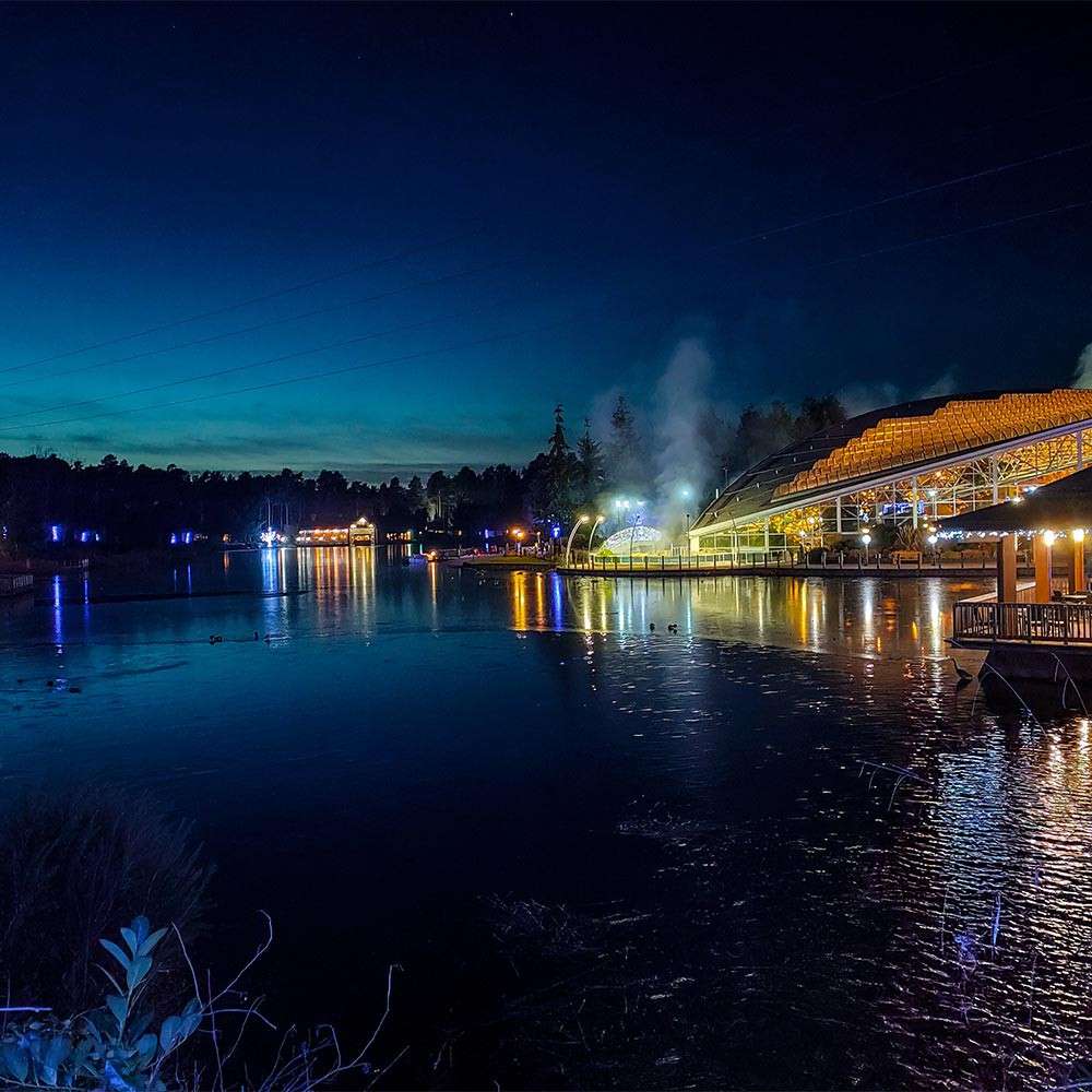 Waterfront buildings glow, casting colored reflections and faint steam onto a calm lake, while ducks drift across; surrounding context features treeline silhouettes, piers, and deep twilight sky.