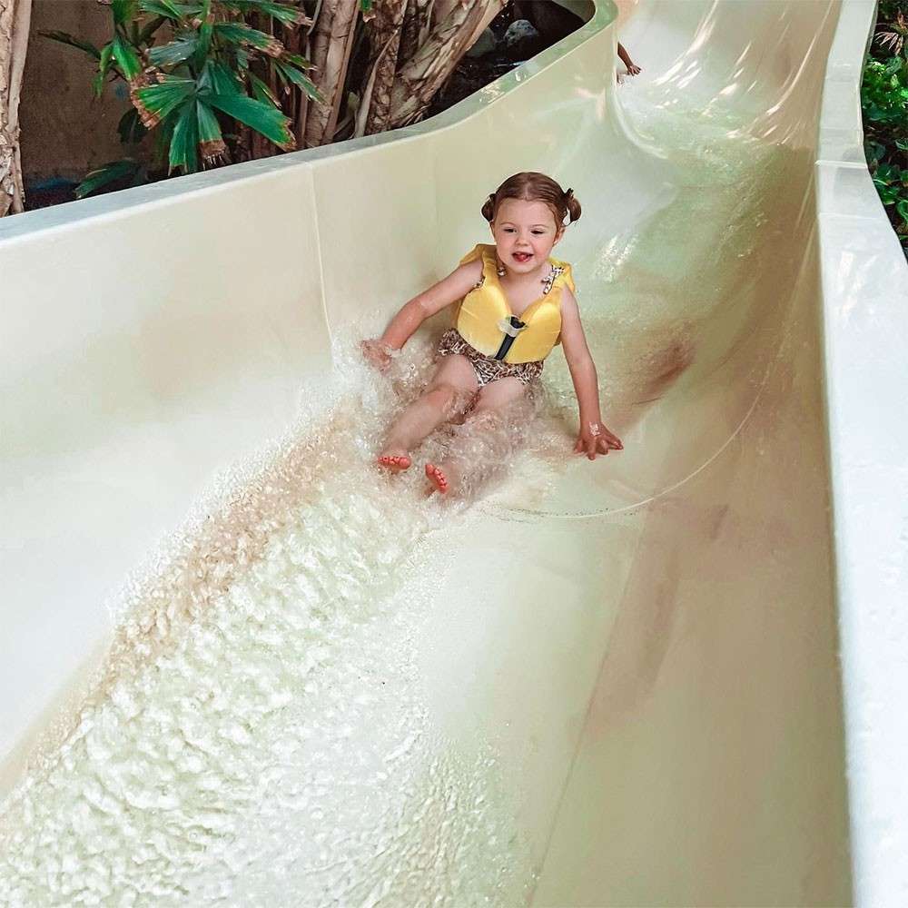 Child wearing a yellow life jacket slides feet-first down a curving water slide, splashing through shallow water, surrounded by tropical plants and cream-colored slide walls in a waterpark setting.
