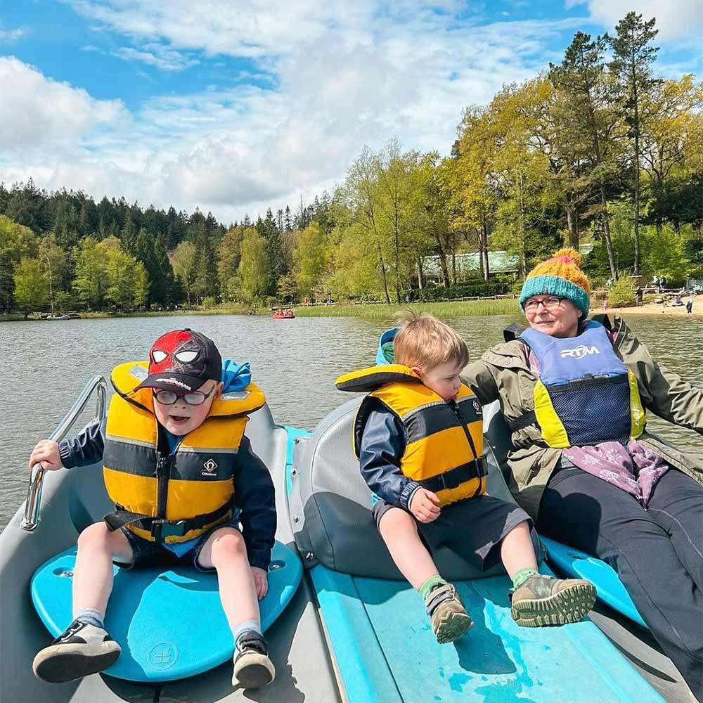 Three people wearing yellow life jackets sit on a blue pedal boat, relaxing, on a calm lake surrounded by trees under a partly cloudy sky. Text: RYA.