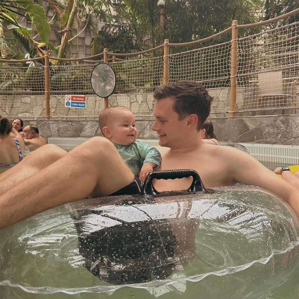 Man and baby lounge in a clear inflatable tube, smiling at each other; indoor waterpark surrounds them with rope fencing, stone walls, and other swimmers. Visible signs: “No diving” and “1.0 m depth.”