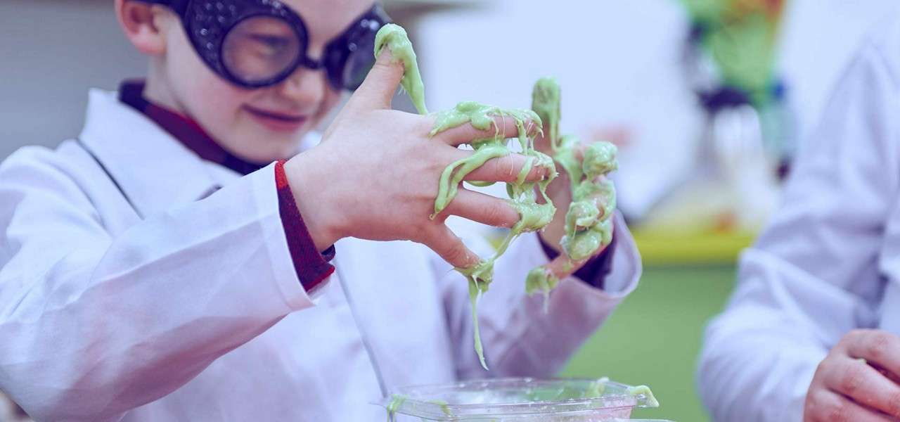 Child in a lab coat and goggles stretches gooey green slime between messy hands, letting it drip into a clear container during a playful science activity in a classroom.