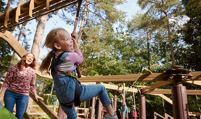 Child rides a zipline handle, laughing, wearing a harness; adult watches smiling behind. Context: wooded adventure playground with elevated rails, posts, and tall trees under a sunny sky.