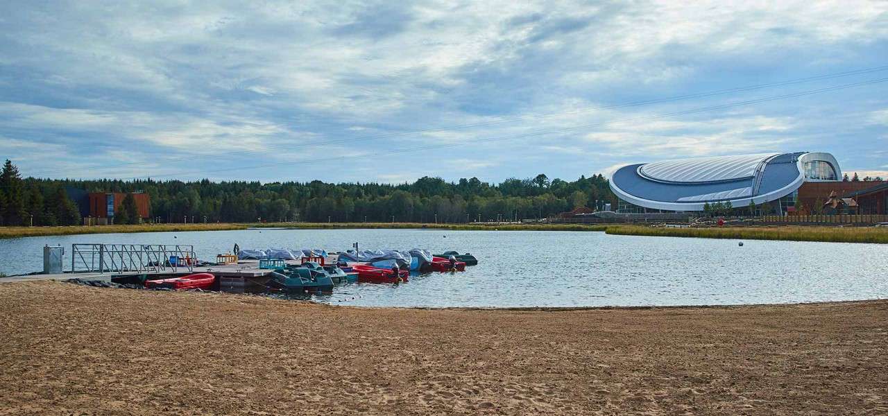 Boats sit moored at a small dock, resting on calm lake water; sandy beach foreground, forested shoreline beyond, and a large futuristic curved-roof building under a cloudy sky.