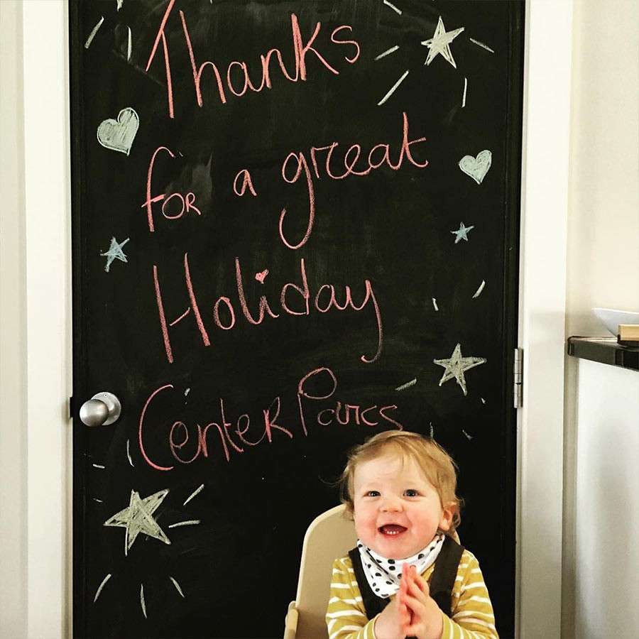 Child smiles and clasps hands while sitting in a high chair, in front of a chalkboard door with stars and hearts. Text: “Thanks for a great Holiday Center Parcs.”