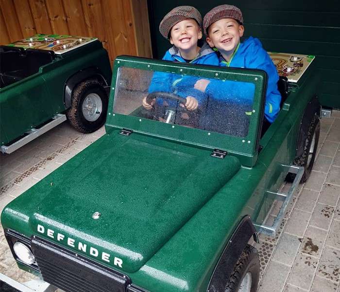 Two children steer and smile in a small green toy jeep labeled DEFENDER, inside a covered, tiled area; another similar vehicle sits nearby.