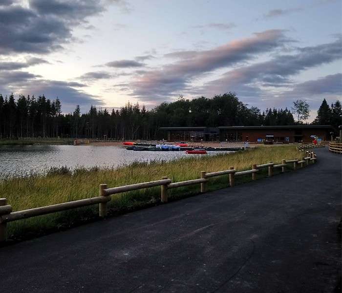 Paddleboats rest at a lakeside dock, evening light reflecting on calm water, while a paved path with wooden railings curves along grassy shore beside a lodge and forested horizon.