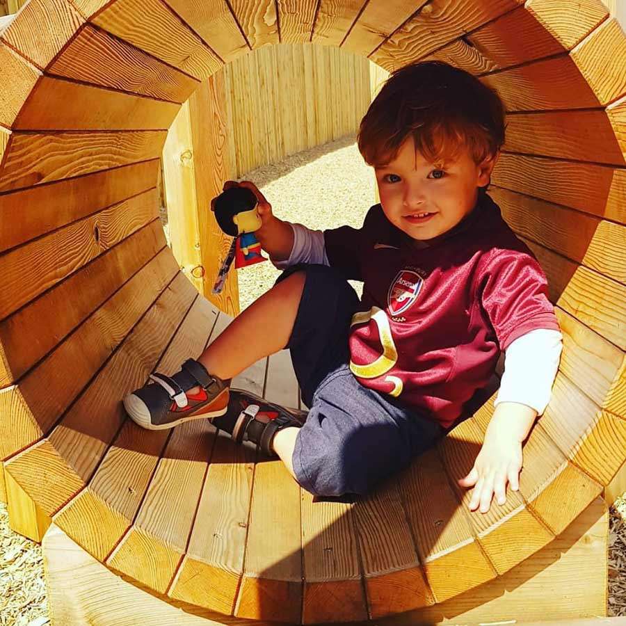 Child sits and smiles, holding a small toy figure, inside a circular wooden tunnel. Sunlit playground surrounds with wood-chip ground and vertical wooden fence in the background.