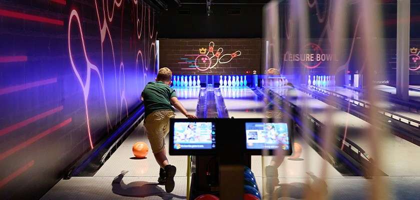 Bowler releases an orange ball; pins glow ahead in a neon-lit alley with digital scoring screens and wall art. Visible text: LEISURE BOWL, 2.